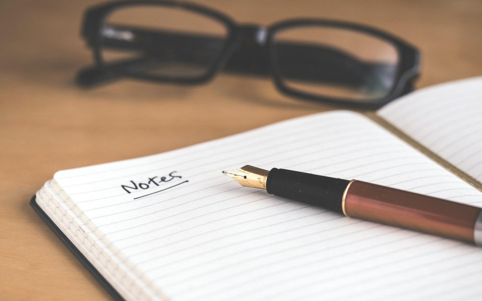Un carnet ouvert, un stylo et des lunettes posés sur un bureau en bois